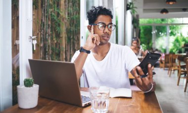 Kafede dizüstü bilgisayar ve kağıt parçalarıyla oturan etnik serbest çalışan ve kafa destekleyen ve akıllı telefon ekranında mesaj okuyan biri.