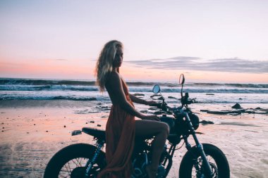 Turuncu ışıklı elbiseli çekici bir kadının yan görünüşü deniz kenarında siyah motosikletle bacaklarını açması nefes kesici su ve gökyüzünün arka planında.