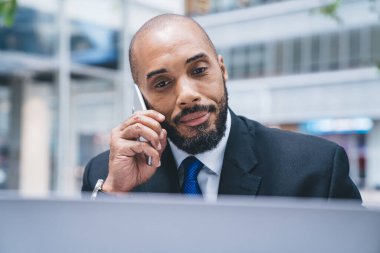 Yakışıklı, kel ve sakallı, takım elbiseli siyahi bir iş adamı ve cep telefonuyla bulanık arka planda dizüstü bilgisayara bakmak. 