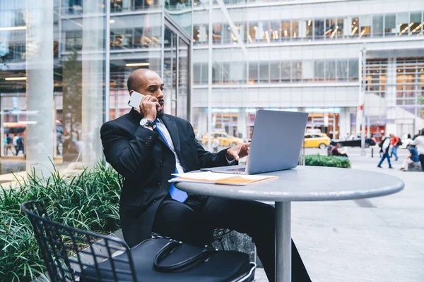 身穿正装的自信的非洲裔美国企业家坐在街上咖啡店的笔记本电脑前 一边用智能手机一边指着屏幕一边说话 — 图库照片