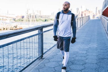 Spor giysili, kel, Afro-Amerikan bir adam. Eldivenleriyle köprüden nehre doğru yürürken etrafına bakıyor.