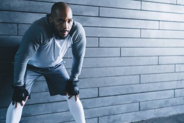 Yakışıklı, Afro-Amerikan, kel, sakallı, spor kıyafetli, elleri kapalı arka planda, dizlerinin üzerinde öne eğilmiş yetişkin bir erkek.