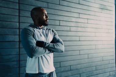 Yakışıklı, dalgın, Afro-Amerikan, kel, sakallı, spor giyimli, kollarını çapraz bağlamış, gri arka plana bakan yetişkin bir erkek.
