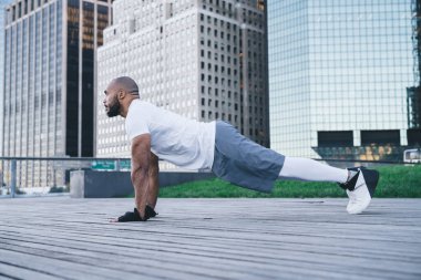 Spor giyimli, kel ve sakallı zenci bir adamın modern şehir merkezinin caddesinde arka planda gökdelenlerle şınav çekerken yan görüntüsü.