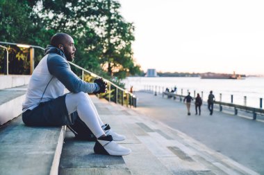 Genç Afro-Amerikan atletik erkek merdivenlerde oturuyor ve şehir caddesine bakarken kulaklıkla müzik dinliyor.