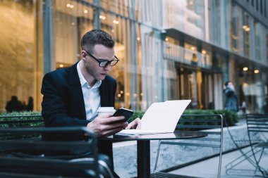 Hırslı erkek yönetici, cam duvarlı binanın bulanık arka planında akıllı telefonu tutarken kaldırım kafesinde oturmuş iş gazetelerini okuyor.