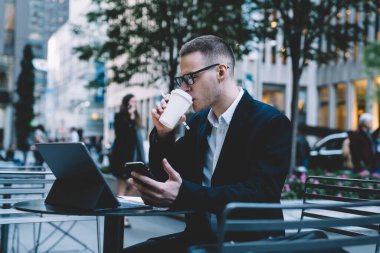 Takım elbiseli ve elinde akıllı telefon olan şık ve yakışıklı bir erkek çalışanın tablet kullanarak tablet tablet içerek sofrada içki içmesinin keyfini çıkarırken görüntüsü 