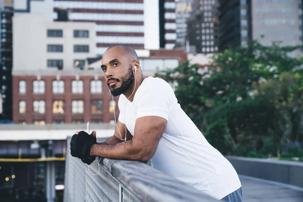 Kenar manzaralı, rüya gibi kel sakallı siyahi adam spor giyim kuşamında gözlerini kaçırıp müzik dinlerken kentteki iskelede korkuluklara yaslanıyor.