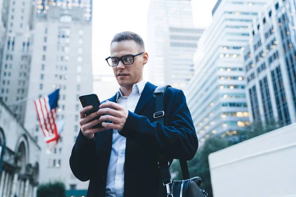 Gözlüklü, kendine güvenen yakışıklı girişimcinin çantasıyla modern şehrin şehir merkezinde bulanık arka planda yürümesinden.