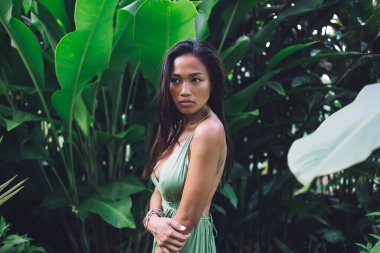 Gerçekten çekici, uzun saçlı, bronzlaşmış tenli, tropikal bahçede ayakta duran, şık yeşil bir elbise giyen Asyalı kadının yan görüntüsü.