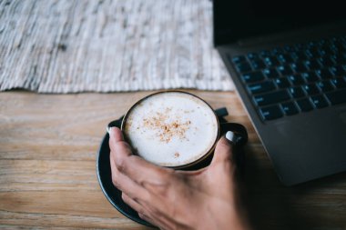 Tanımlanamayan bir kadının elinde kapuçinolu kahve fincanı, ahşap masa örtüsü, gri masa örtüsü ve bulanık dizüstü bilgisayarla kapuçino tutması.