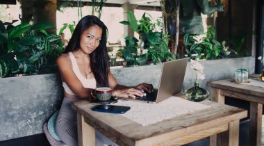 Eğik kafalı, esrarengiz bir gülümsemesi olan genç ve güzel bir bayan bir fincan çay ve akıllı telefonla masada oturuyor ve kameraya bakarken dizüstü bilgisayarda çalışıyor.