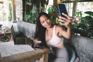 Egzotik bir kafede oturup laptopla kahve içen, gülümseyen ve akıllı telefonuyla pohpohlayan güzel etnik bayan.