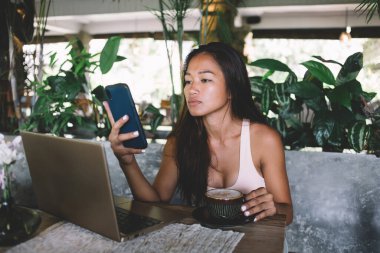 Bilgisayarlı, kahveli ve akıllı telefondan haber okuyan kendine güvenen Asyalı bir kadın.
