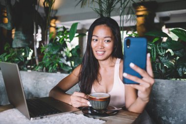 Partnerle iş birliği projesini tartışan genç serbest çalışan güneşli bir günde rahat kafeteryada cep telefonu ve kapuçino içerken görüntülü sohbete yardım ediyor.