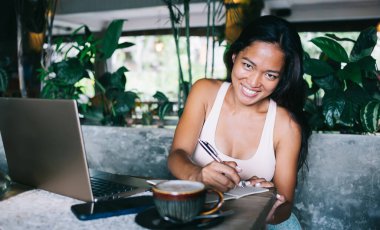 Gülümseyen, neşeli, seksi flört eden, gülen, beyaz dişli Asyalı kadın not defterine notlar alıyor ve masa dolusu dizüstü bilgisayar, akıllı telefon ve kahve dolu.