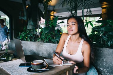 Genç bir kadının kapuçino ve dizüstü bilgisayarla masada oturduğunu ve not defterine not aldığını ve kafeye bakmadığını hayal ediyor.