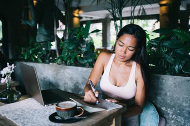 Asyalı, düşünceli genç bayan kafeterya masasında dizüstü bilgisayarla çalışırken yeni dizeler yazıyor.