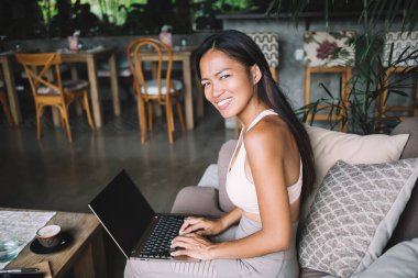 Bilgisayarda yazı yazarken ve kafede rahat bir kanepede otururken kameraya bakan asabi genç bir kadının yan görüntüsü.