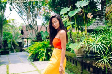 Genç, rüya gibi, uzun saçlı, etnik bir bayanın tropikal parkta yürürken kameraya bakışının yan görüntüsü.