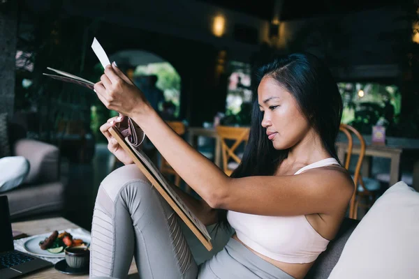 Beyaz ve gri spor kıyafetleri giymiş rahat gülümseyen bir kadının yan görüntüsü. Gri kanepede oturmuş menülerin sayfalarını çeviriyor.