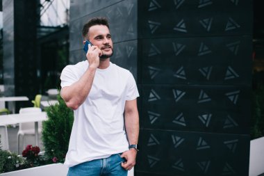 Gülümseyen sakallı bir erkek, cebinde gündelik giysiler, akıllı telefonuyla konuşurken, yaratıcı binanın dış cephesinde dikilip başka yöne bakıyor.