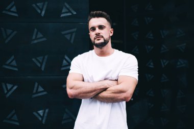 Çapraz kaslı kolları olan, modern ofis binasının önünde dikilirken kameraya bakan ciddi bir erkek.