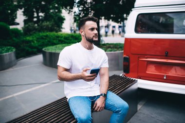 Parkta bankta oturan, akıllı telefon ve kulaklıkla müzik dinleyen, sıradan giyinen yakışıklı bir adam.