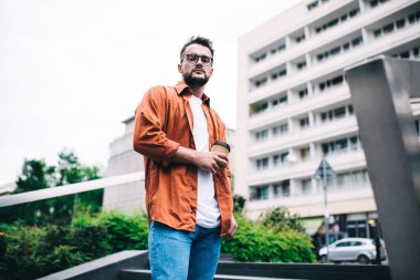 Gözlüklü ve günlük kıyafetleri olan kendine güvenen bir adam park yerinde durup dinlenirken mola sırasında kahvenin tadını çıkarıyor ve kameraya bakıyor.