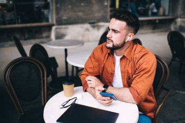 Düşünceli, yakışıklı, sakallı, serbest çalışan, kablosuz kulaklıklı müzik dinleyen, akıllı telefon kullanan, kafede oturmuş, gözlerini başka yöne çeviren biri.