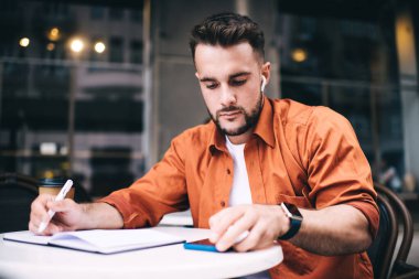 Kablosuz kulaklıklar takan, günlük turuncu gömlekli, akıllı telefonlu, yakışıklı, sakallı bir adam kafede otururken hatırlatıcı notlar alıyor.