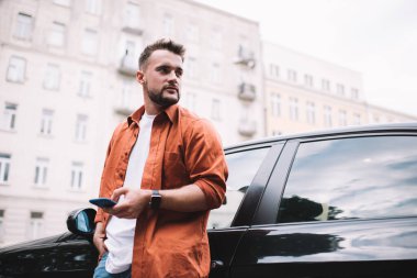 Düşük açılı turuncu tişörtlü, kendine güvenen erkek. Elini cebinde tutuyor ve gözünü kaçırıyor. Sokakta siyah arabanın yanında akıllı telefon kullanıyor.