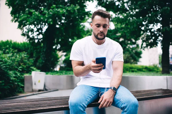 スマートフォンでスマートウォッチメッセージを発信し ベンチに座っている間にヘッドフォンで音楽を聴くカジュアルな服で髭を生やした男性 — ストック写真