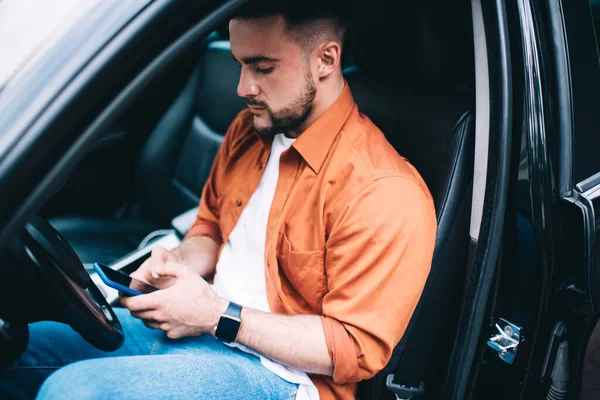 Sıradan giysiler içinde sakallı, camları açık arabada oturan ve modern akıllı telefonu dikkatle kullanan düşünceli bir erkek sürücü.