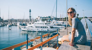 Barcelona şehrinin limanında yatların yanında otururken kameraya bakıp müzik dinleyen, güneş gözlüklü genç sarışın bir kadın.