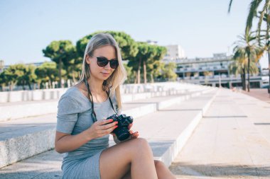Güneşli yaz günlerinde Barselona 'da merdivenlerde otururken modern kamerada fotoğraf kontrol eden güneş gözlüklü sarışın kadın.