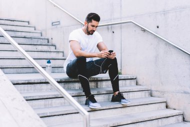 20 'li yaşlardaki kendinden emin erkek aktif giyimde caddede duran akıllı telefondan bildirimleri kontrol ediyor, ciddi erkek blogcu takipçileriyle sağlıklı yaşam tarzı medya ve müzik paylaşımı hakkında iletişim kuruyor.