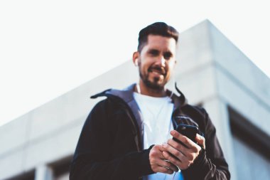 Adamın elindeki modern akıllı telefona odaklanmak, aktif ceketli ve kulaklıklı gülümseyen beyaz hippi adamın sağlıklı yaşam tarzını ve eğitimini sürdürmek için aletler kullanmaktan memnun olduğu bulanık görüntüsü.