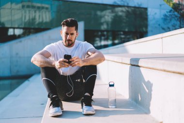 Aktif haldeki bin yıllık hipster çocuğu cep telefonundan spor programı indirdikten sonra test sonuçlarını kontrol ediyor. Akıllı telefon ve kulaklık kullanarak antrenmandan sonra dinlenen sakallı genç bir sporcu.