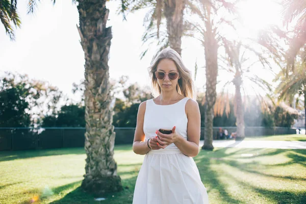 公園の芝生の上に立つ白いドレスとサングラスの若いブロンドの女性とバルセロナ市内の晴れた日にスマートフォンを使用して — ストック写真
