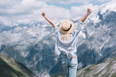 Sıradan giyinmiş, şapkalı, yumruklarını sıkan ve başarıyı kutlayan bir bayanın, Lago di Braies gezisi sırasında dağ sırasının karşısında duruşu.