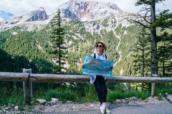 Happy young woman with map smiling and looking away while sitting on fence against mountain ridge during trip to Karersee Lake