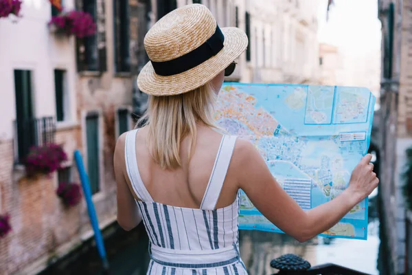 帽子で若いブロンドの女性観光客のバックビュー地図を見て イタリアのヴェネツィアの通りに立っている間場所を確認 — ストック写真