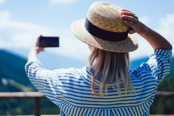 在阳光明媚的日子里 年轻女子一边触摸帽子 一边拿着智能手机自拍的倒影 — 图库照片