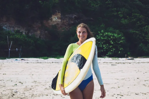 快乐而迷人的年轻女子 身穿泳衣 一边微笑着 一边享受假期 一边手握冲浪板 站在阳光灿烂的海滩上 — 图库照片