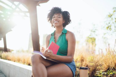 Neşeli esmer tenli kadın, şehir parkında boş zamanlarını değerlendiriyor hafta sonları günlük planlama notları alıyor, mutlu Afro-Amerikan kadın blogcu, güneşli bir günde eğlenmek için yayın için fikirler yazıyor.
