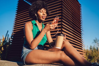 Kaygısız siyah kadın, güneşli bir günde parkta oturmuş kahve içerken müziğin keyfini çıkarıyor ve akıllı telefondan şarkı seçiyor.