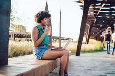 Gülümseyen kıvırcık saçlı, yeşil tişörtlü, şortlu, laptopta sörf yapan ve yazın kahve içen bir kadın. 