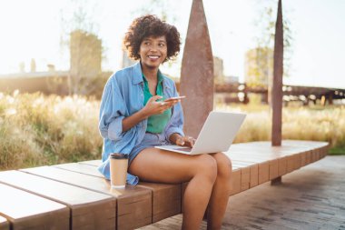 Yeşil gömlekli, kot pantolonlu, kıvırcık saçlı, mutlu bir etnik bayan. Cep telefonu ve dizüstü bilgisayarla yazın sokakta bir fincan kahveyle oturuyor. 