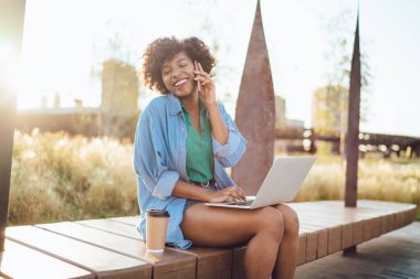 Yeşil tişörtlü ve şortlu mutlu etnik bayan cep telefonuyla sohbet ediyor. Yazın bir fincan kahve içip dizüstü bilgisayarla sokakta sörf yapıyor. 